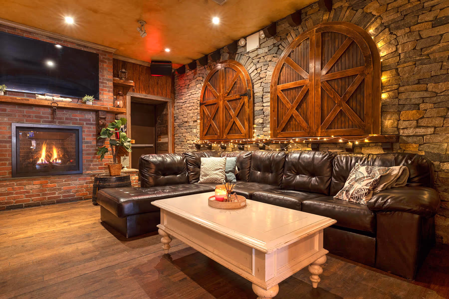Cozy lounge with dark leather sectional sofa, white coffee table, lit fireplace, and stone walls decorated with wooden shutters and string lights.