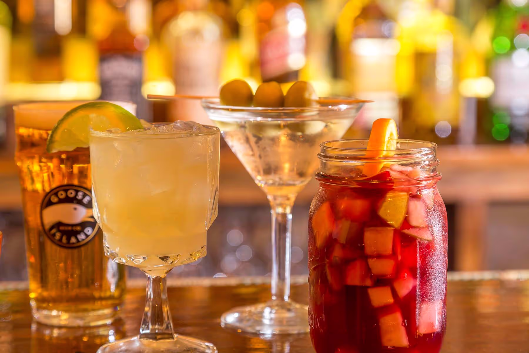Four colorful cocktails on a bar counter with a blurred background of liquor bottles.