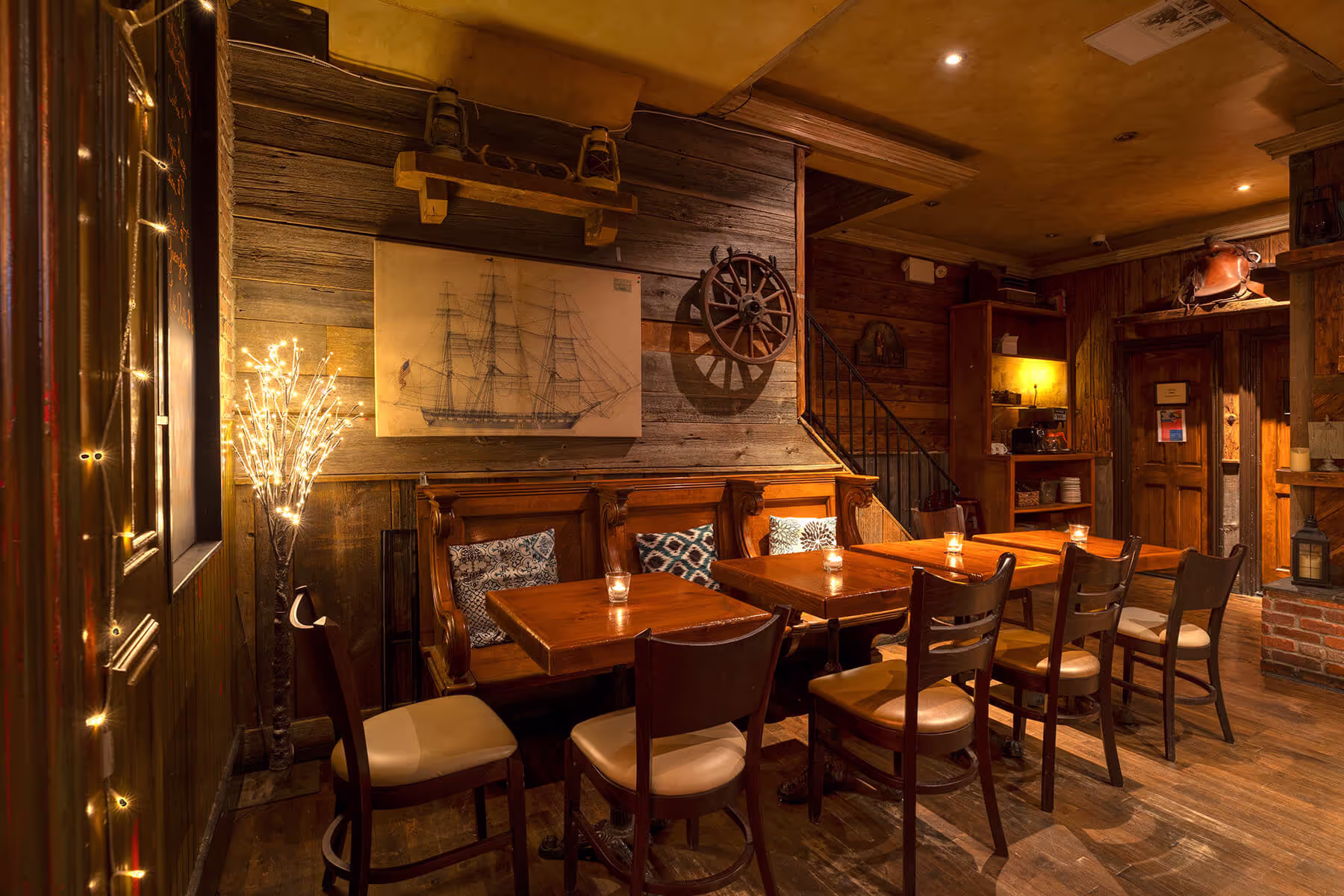 Cozy wooden interior of a rustic restaurant with tables, chairs, cushions, warm lighting, and nautical decor including a ship painting and a ship wheel.