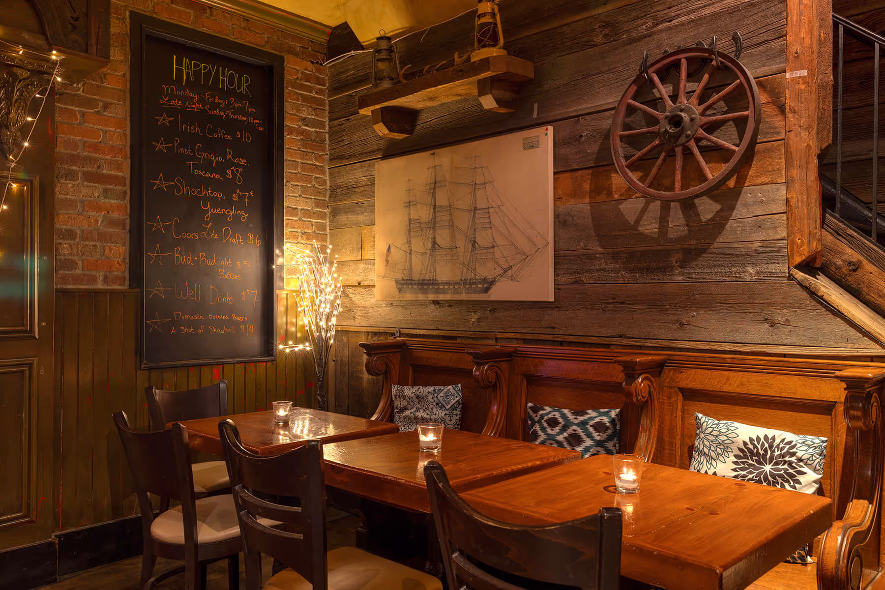 Cozy bar corner with wooden tables, chairs, cushioned bench seating, candles, and a blackboard listing happy hour drinks under warm lighting.