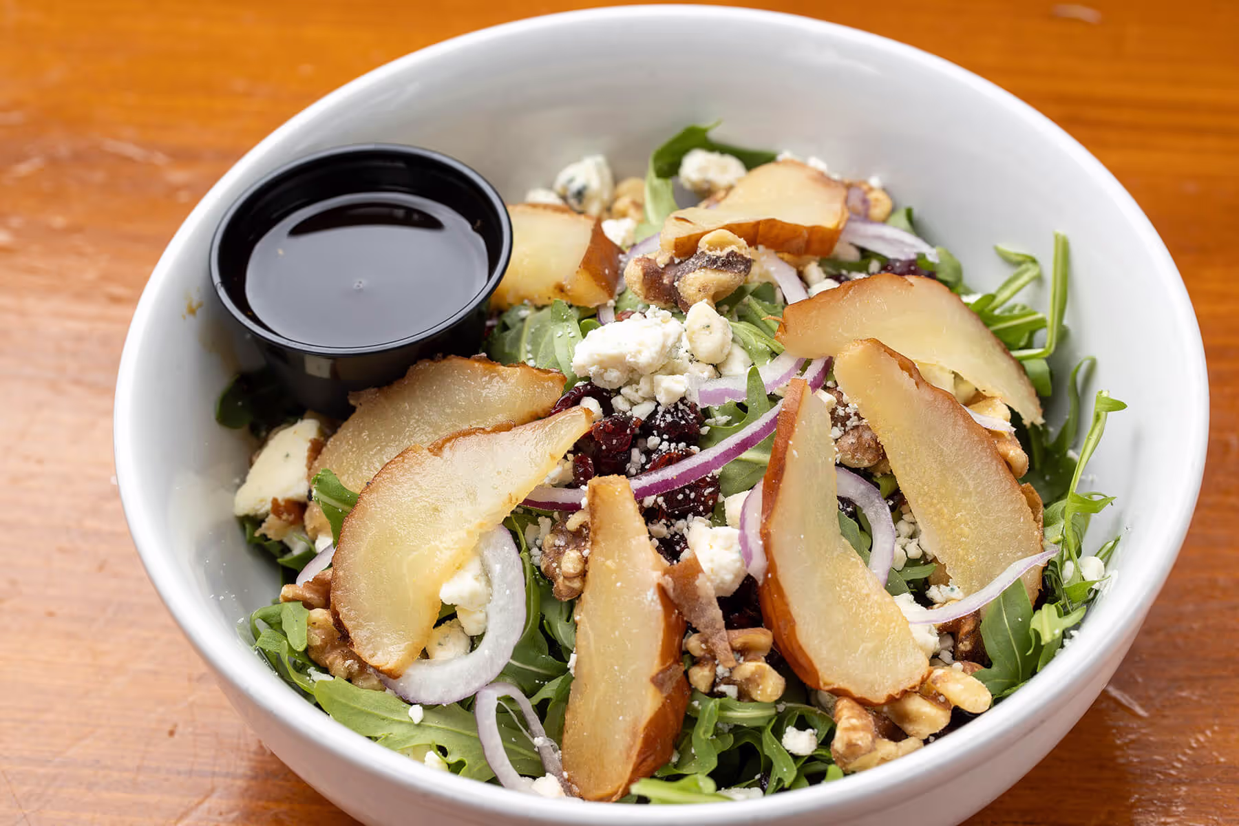 White bowl of arugula salad with sliced pears, walnuts, red onion, blue cheese crumbles, dried cranberries, and a small container of dressing.