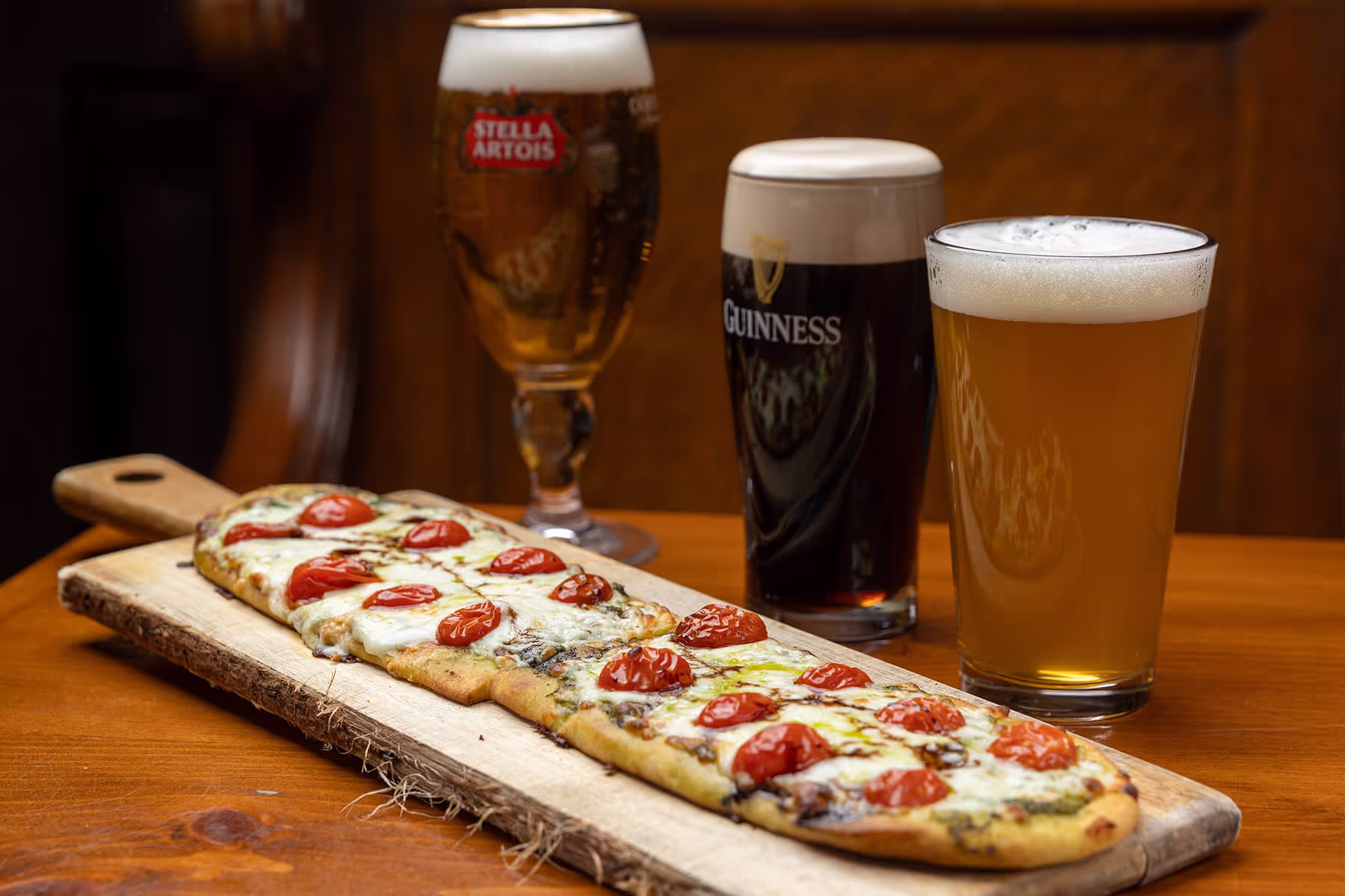 Flatbread pizza with cherry tomatoes and mozzarella cheese on a wooden board, accompanied by three glasses of beer including Stella Artois and Guinness.