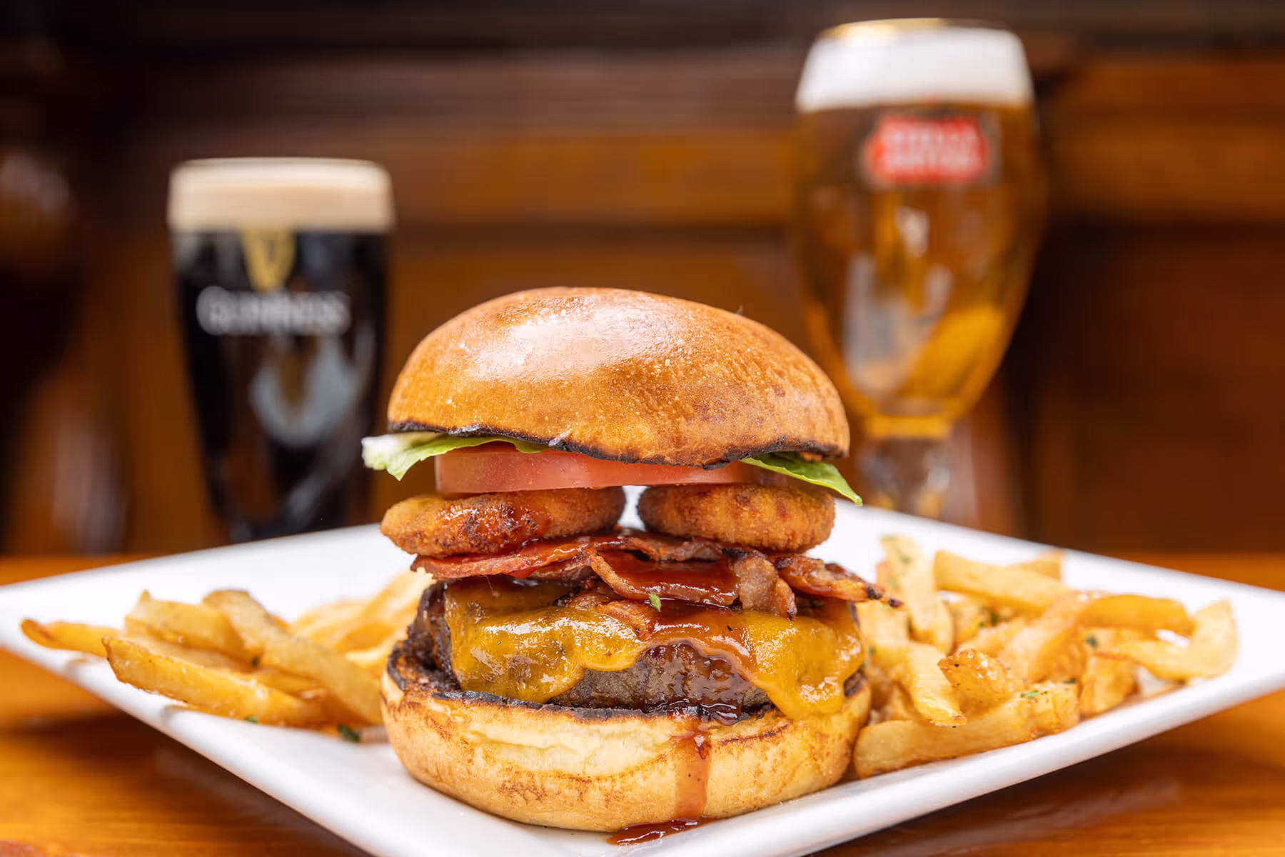Cheeseburger with bacon, fried onion rings, tomato, and lettuce, served with French fries and two glasses of beer in the background.