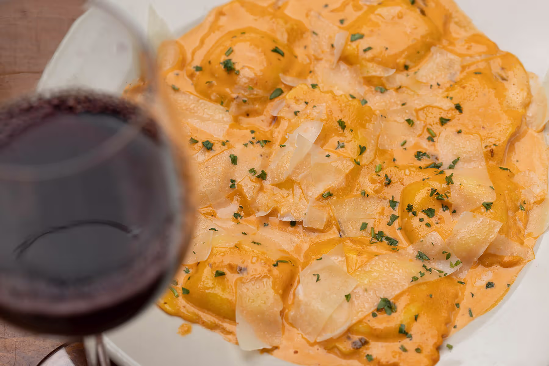 Plate of ravioli pasta in creamy orange sauce garnished with shaved Parmesan and chopped parsley, with a glass of red wine in the foreground.