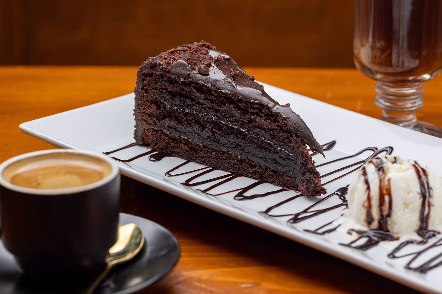 Slice of layered chocolate cake with chocolate drizzle on a white plate, served with a scoop of vanilla ice cream drizzled with chocolate sauce, and a cup of coffee.