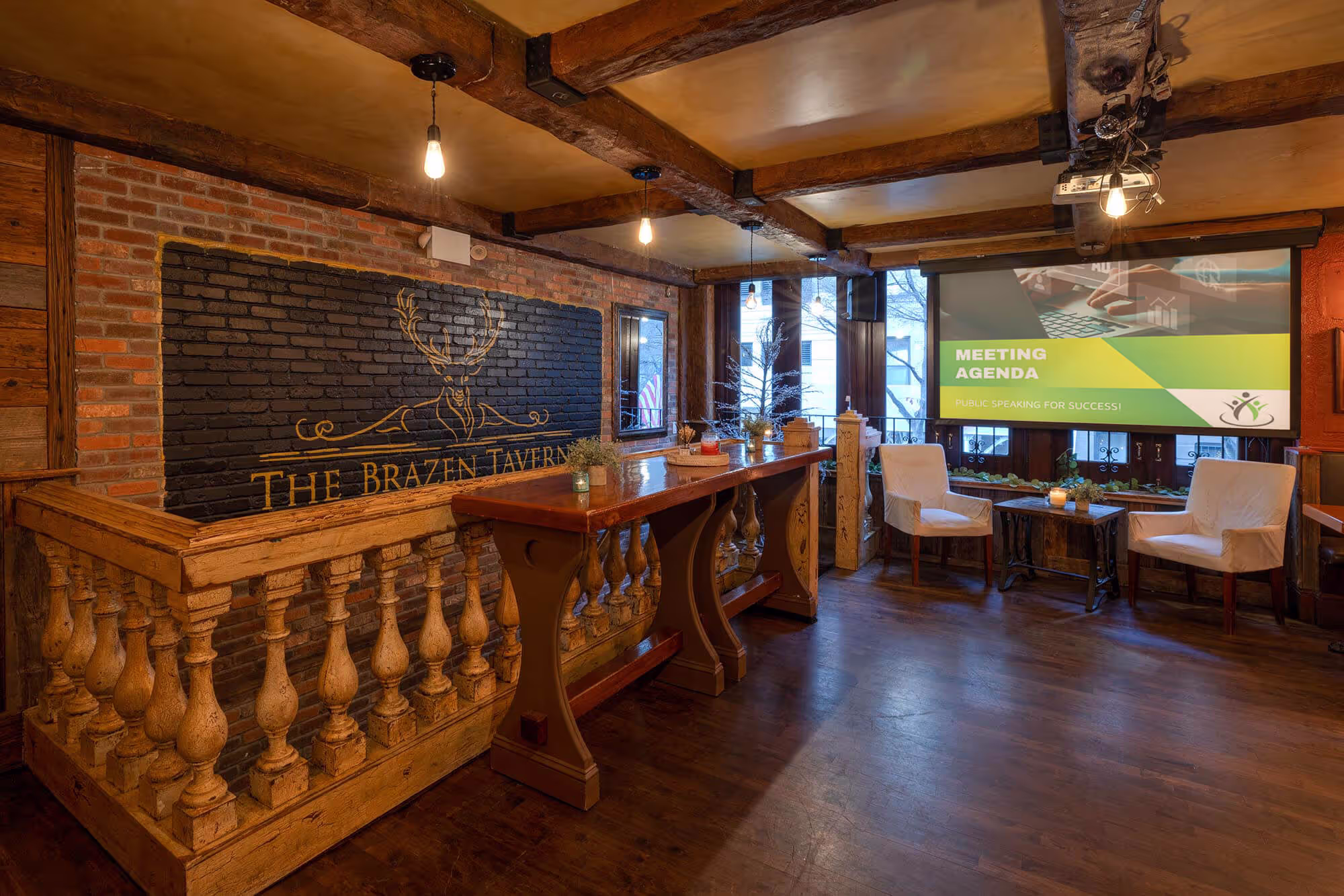Rustic meeting room with wooden beams, two white chairs, a high table, and a screen displaying 'Meeting Agenda: Public Speaking for Success!'