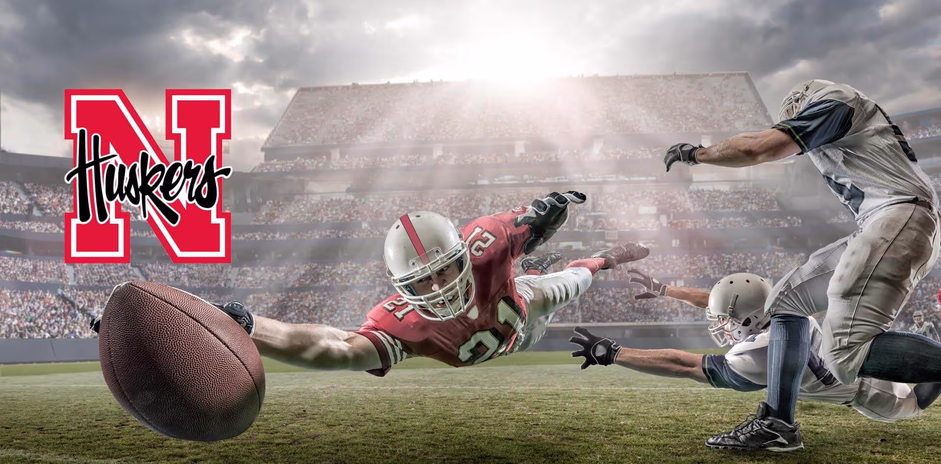 American football player in red uniform diving to reach football near two defenders in white uniforms on a stadium field with Nebraska Huskers logo on the left.