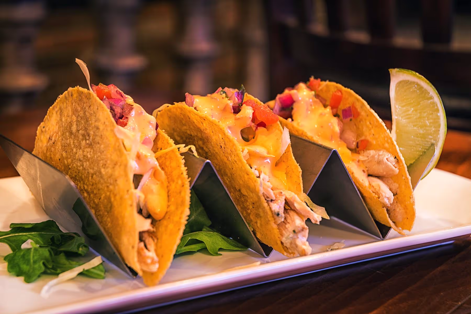 Three tacos filled with chicken, cheese sauce, diced tomatoes, and red onions, served with a lime wedge on a white plate.