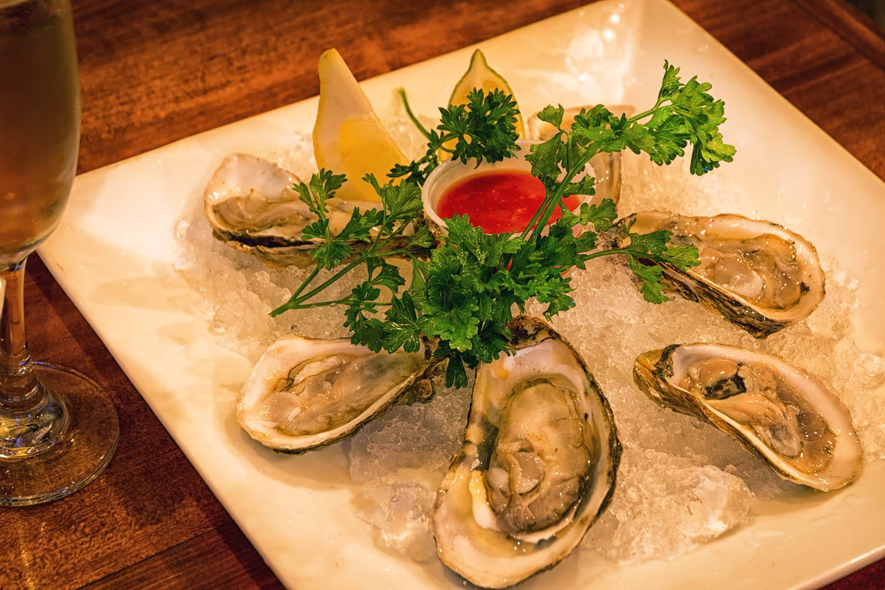 Oysters served on crushed ice with lemon wedges, parsley garnish, and a cup of cocktail sauce on a white square plate.