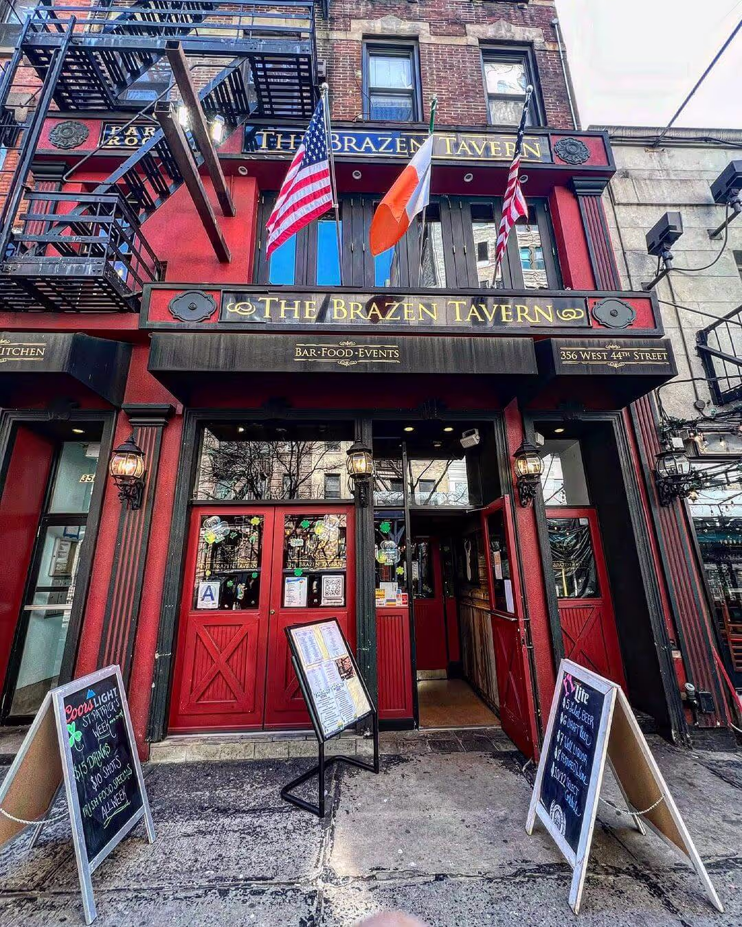 Front entrance of The Brazen Tavern with red doors, American and Irish flags, and outdoor menus on a city sidewalk.