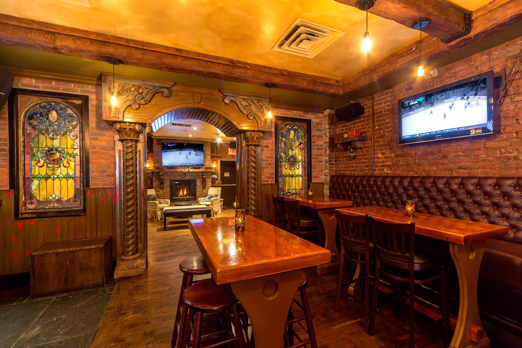 Cozy bar interior with wooden tables, tufted leather seating, exposed brick walls, stained glass windows, and TVs showing a hockey game.